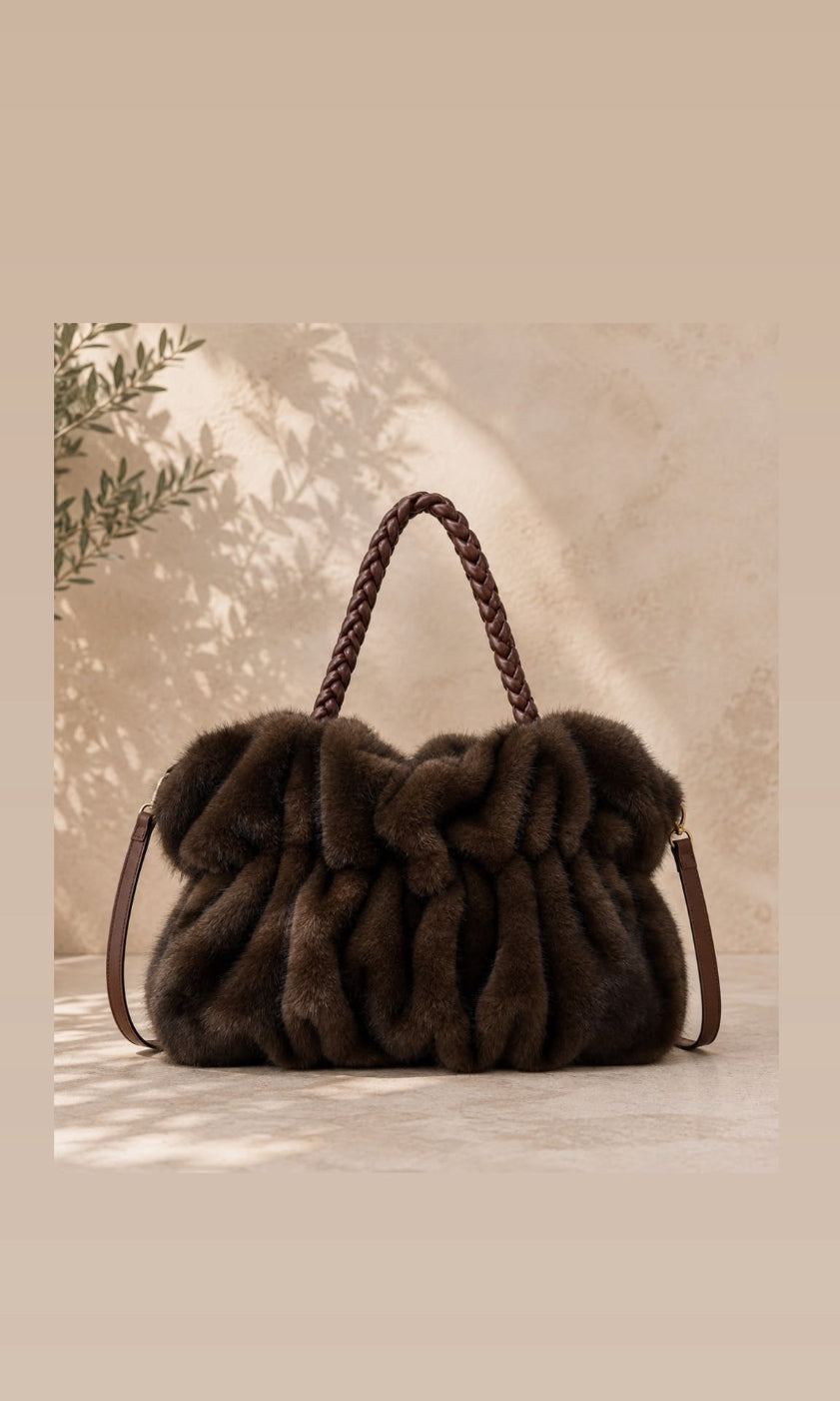 A dark brown plush fur shoulder bag with braided leather handles and a thin shoulder strap, displayed against a warm beige textured background with soft leaf shadows.