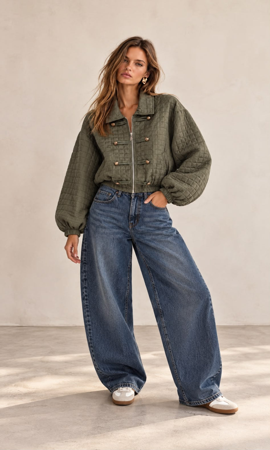 A woman modeling a cropped, quilted olive green jacket with wide-leg blue jeans and white sneakers, standing against a neutral beige studio backdrop.